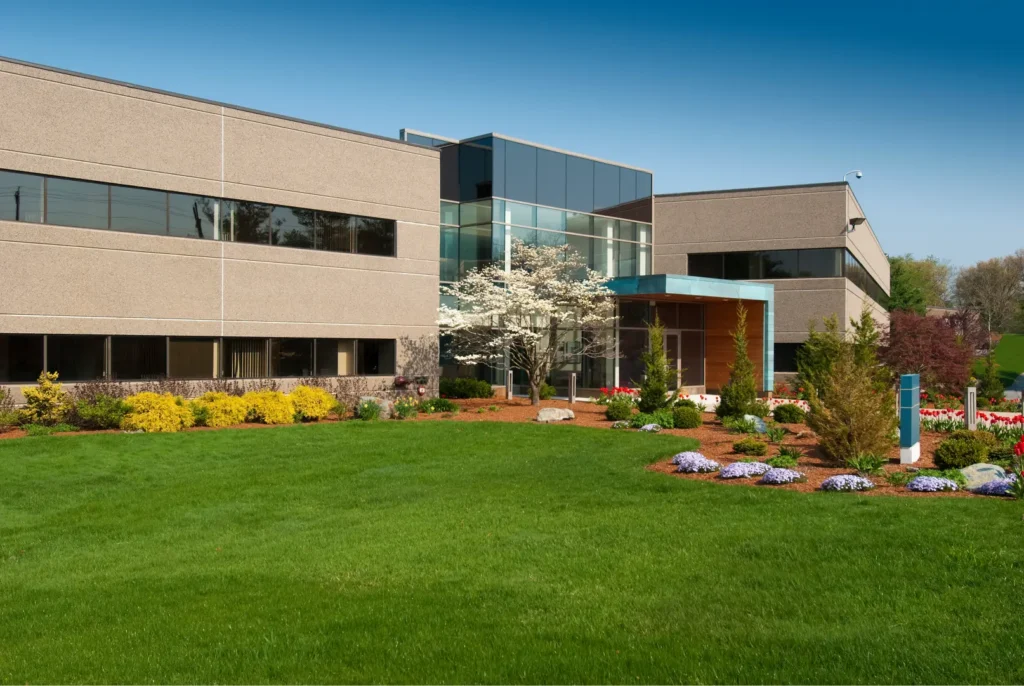 Modern office building with large glass entrance and landscaped garden with green lawn and flowering plants.