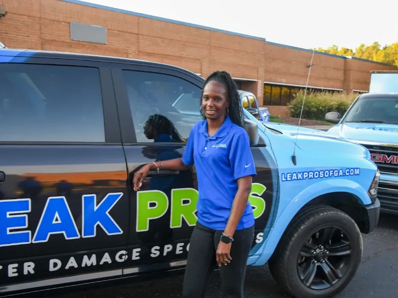 Woman in a blue polo shirt standing next to a black and blue vehicle with "Leak Pros" signage.