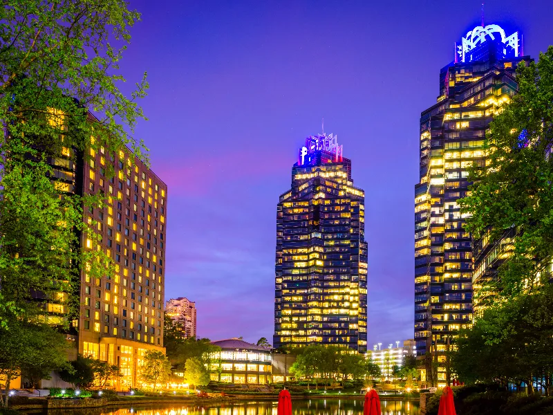 Two illuminated skyscrapers and a hotel building reflected in a river at dusk.