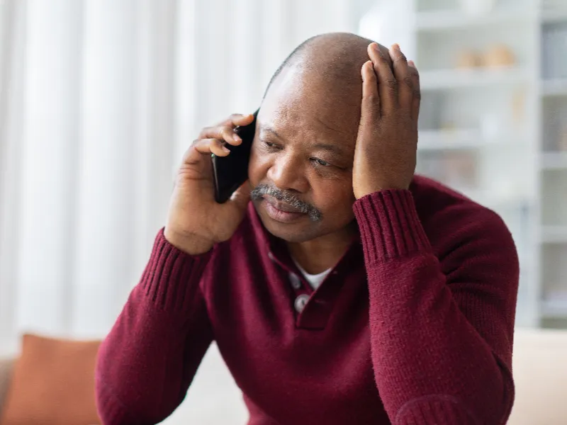 A man in a maroon sweater looks worried while talking on a phone indoors.
