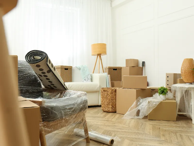 Living room with packed cardboard boxes, wrapped furniture, and a rolled-up rug ready for moving.