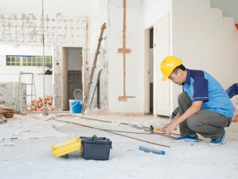 Construction worker wearing a yellow hard hat using a hammer on a concrete floor inside a building under renovation.