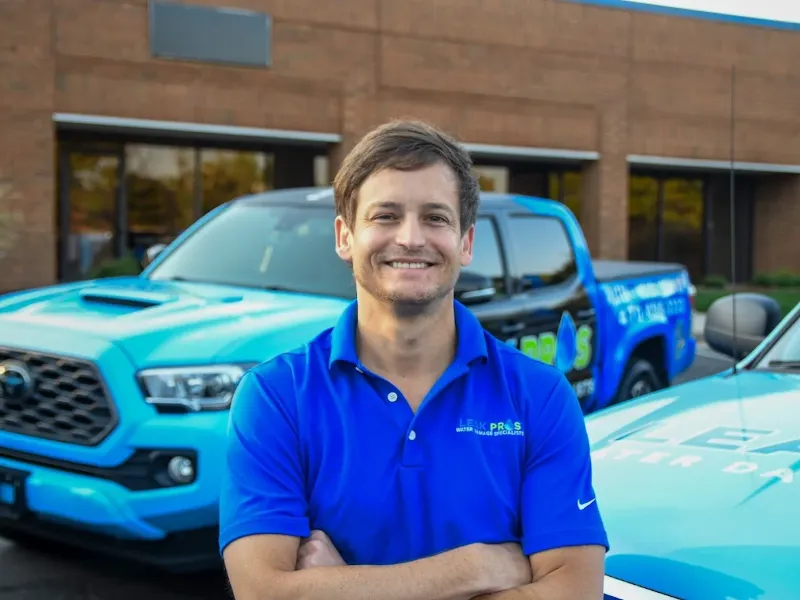 Man in a blue polo shirt smiling with arms crossed in front of blue company trucks.