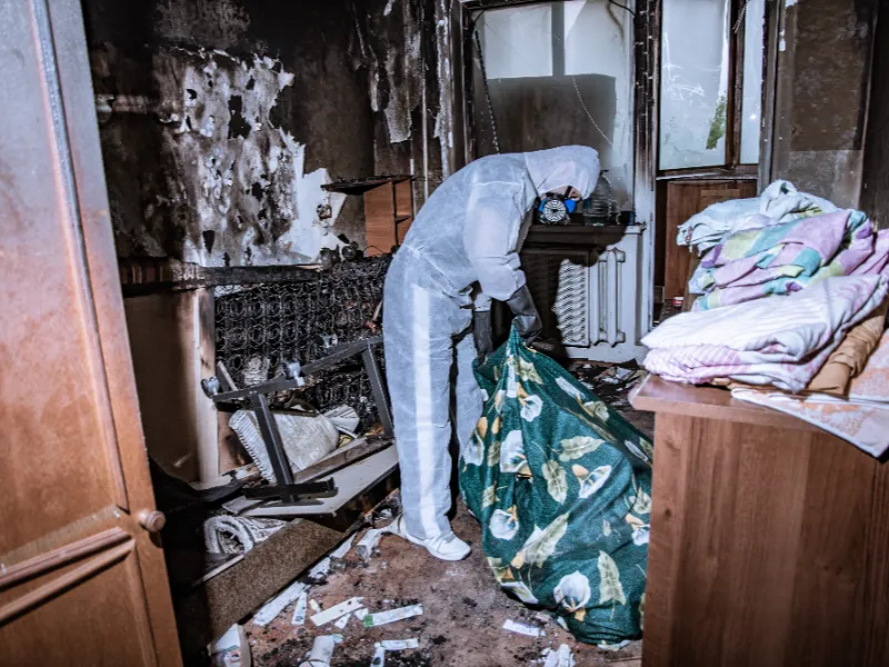 Person in protective suit and mask handling debris in a fire-damaged room.