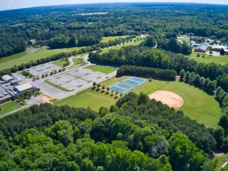 Aerial view of a sports complex with tennis courts, a baseball field, parking lots, and surrounding trees.