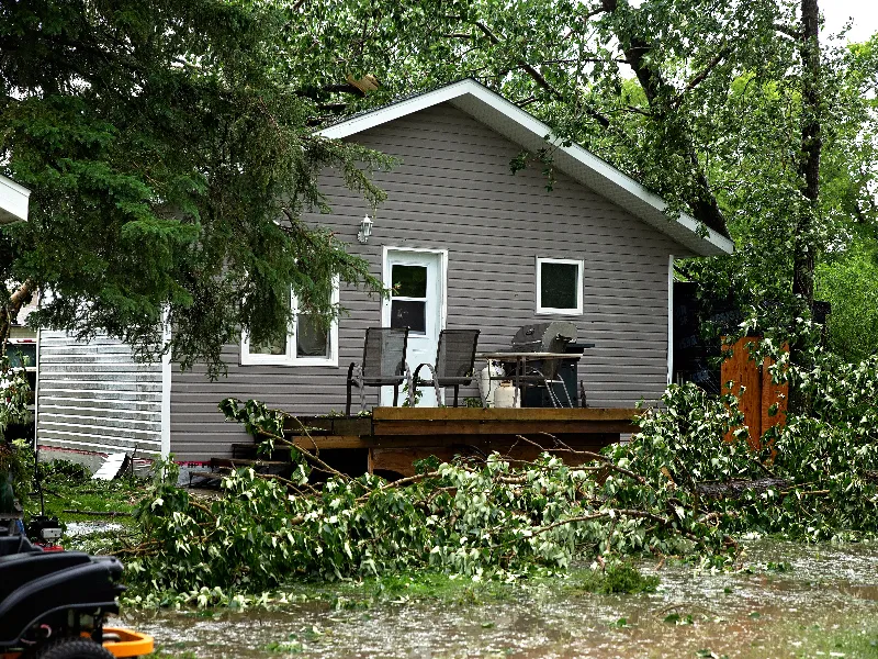 Flooded yard with fallen tree branches in front of a gray house with a wooden deck and outdoor chairs.