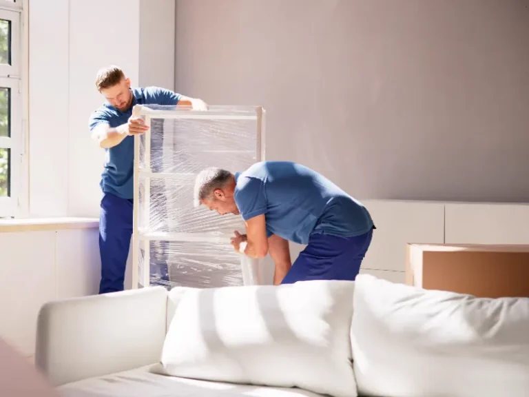 Two men in blue shirts wrapping a large framed object in plastic wrap inside a room.